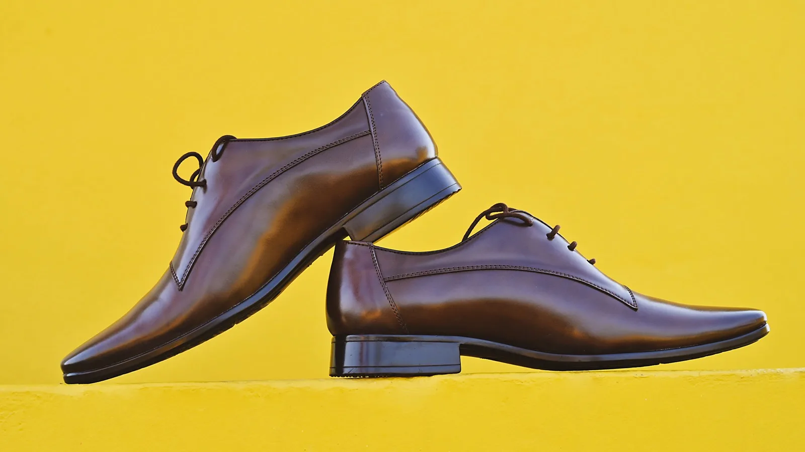 Two brown leather shoes photographed against a bright yellow wall